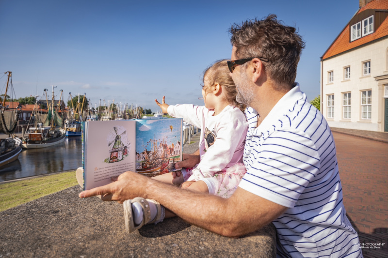 Ein Vater ist mit seiner Tochter an der Hafenmauer in Greetsiel mit dem geöffneten Buch "Deichkrönchen" in der Hand. Beide schauen auf die Kutter im Hafen.
