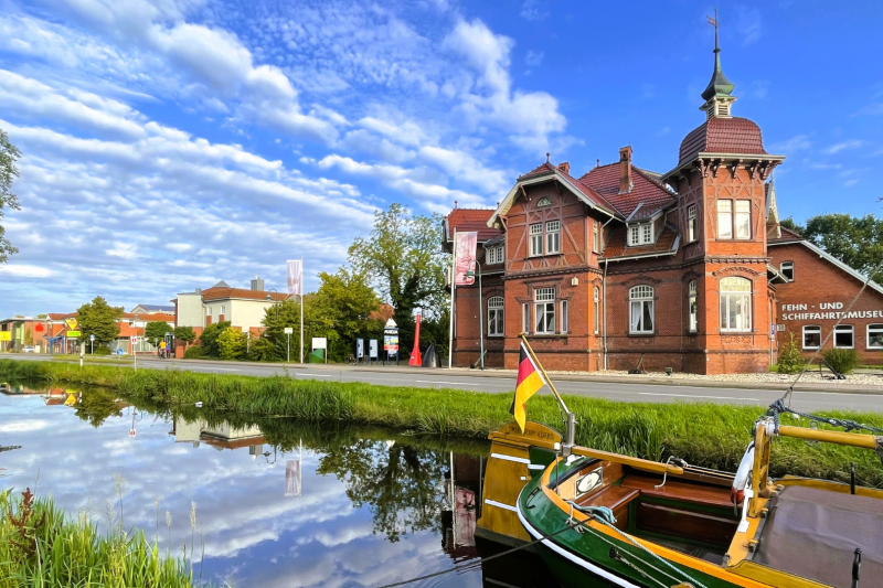 Das Fehn- und Schiffahrtsmuseum in Westrhauderfehn mit einem grünen Plattbodenschiff im Vordergrund auf dem Wasser und Schäfchenwolken am blauen Himmel.