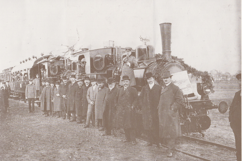Gruppe von Männern in Mänteln und Hüten steht vor einer historischen Dampflokomotive mit angehängten Waggons auf Bahngleis