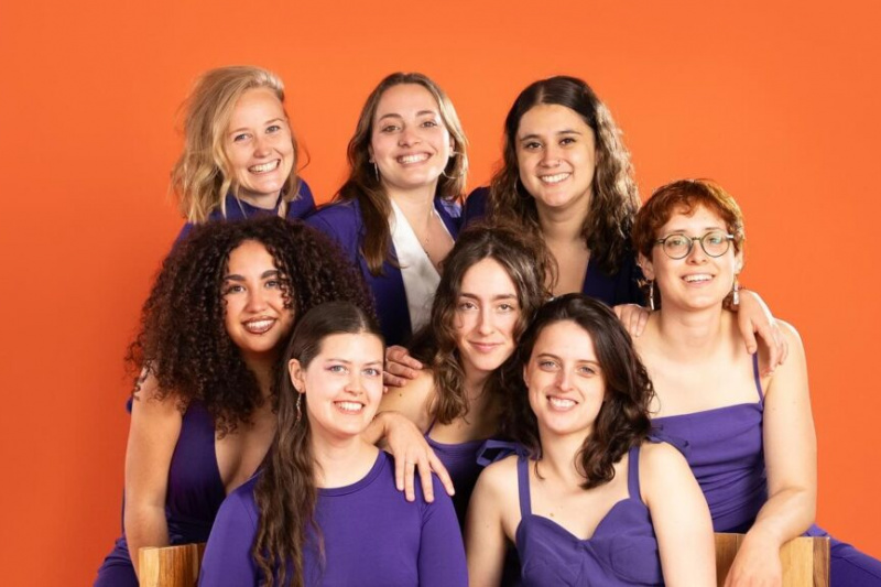 Acht junge Frauen in lila Outfits lächeln vor einem leuchtend orangen Hintergrund.