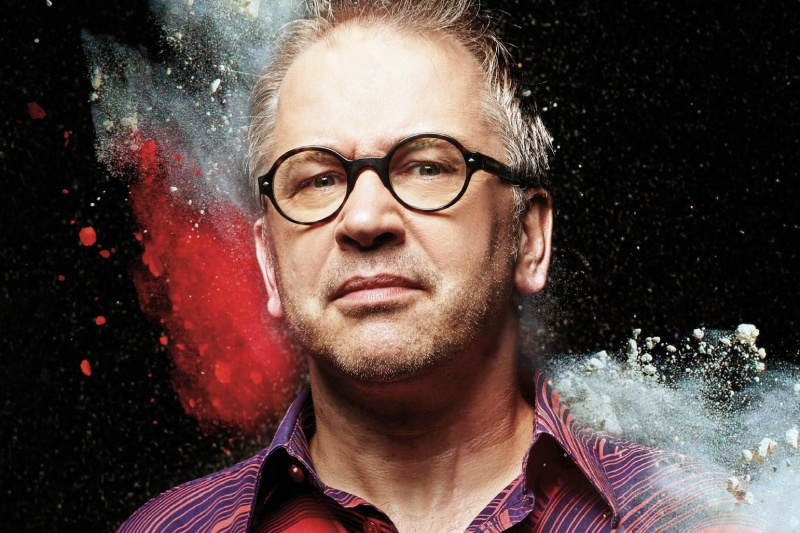 Porträt von Robert Griess mit Brille und buntem Hemd. Im Hintergrund explodiert farbiges Pulver.