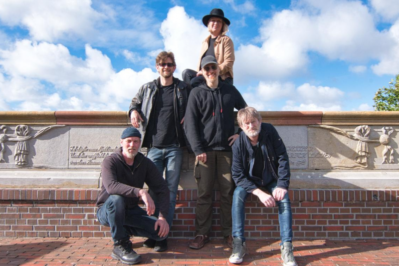 Die fünfköpfige Band Van't Siel posiert draußen vor einer Steinmauer unter blauem Himmel.