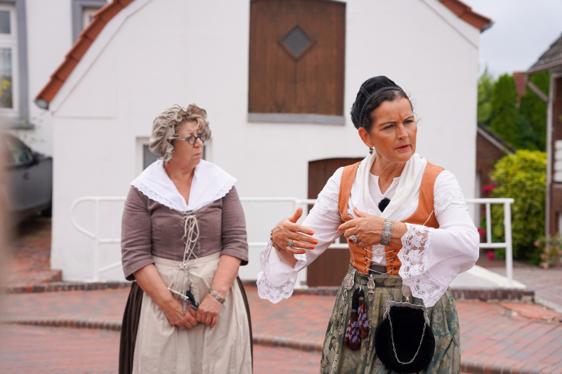 Zwei Frauen in historischen Kostümen führen durch Greetsiel vor einem weißen Haus.