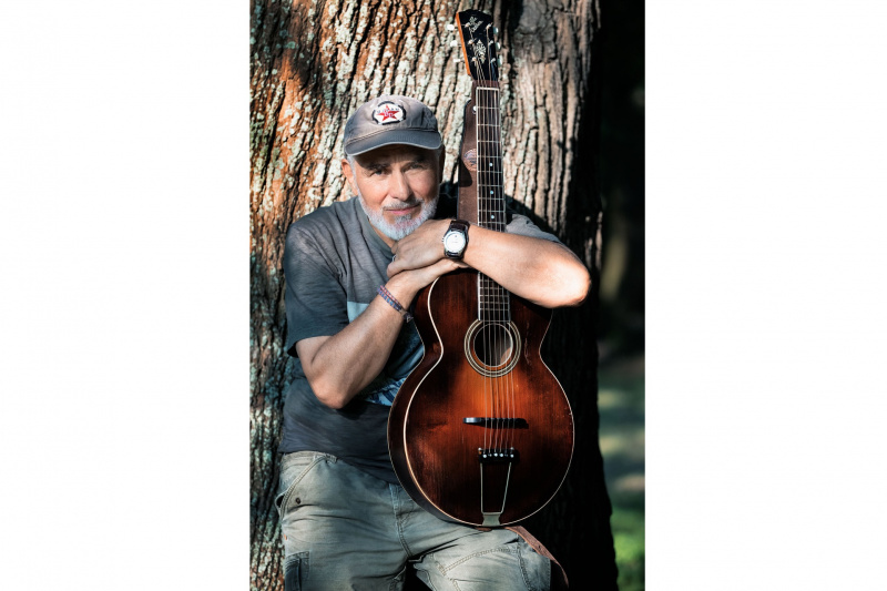 Hansa mit Akustikgitarre vor einem Baumstamm im Freien.