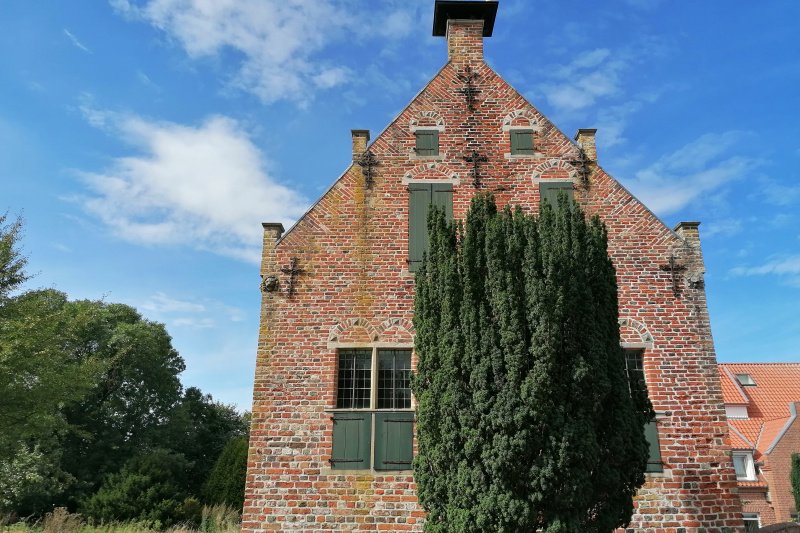 Backsteinbau mit grünen Fensterläden und markanter Giebelfront, teils verdeckt durch hohen Baum.