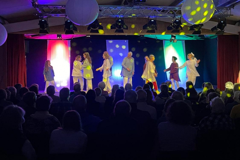 Foto einer Aufführung von "De lüttje Dag", auf der Bühne sieht man die Darstellenden tanzen, im Bühnenhintergrund sieht man drei bunt beleuchtete Vorhänge. Bunte Lichtpunkte tanzen über das Bühnenbild.