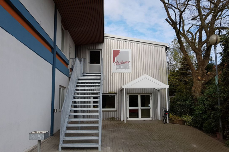 Foto, das den Haupteingang des Haxtumer Speichers zeigt. Eine Metalltreppe führt zur Eingangstür, neben der auf einem großen Schild das Logo des NDT Aurich zu sehen ist.