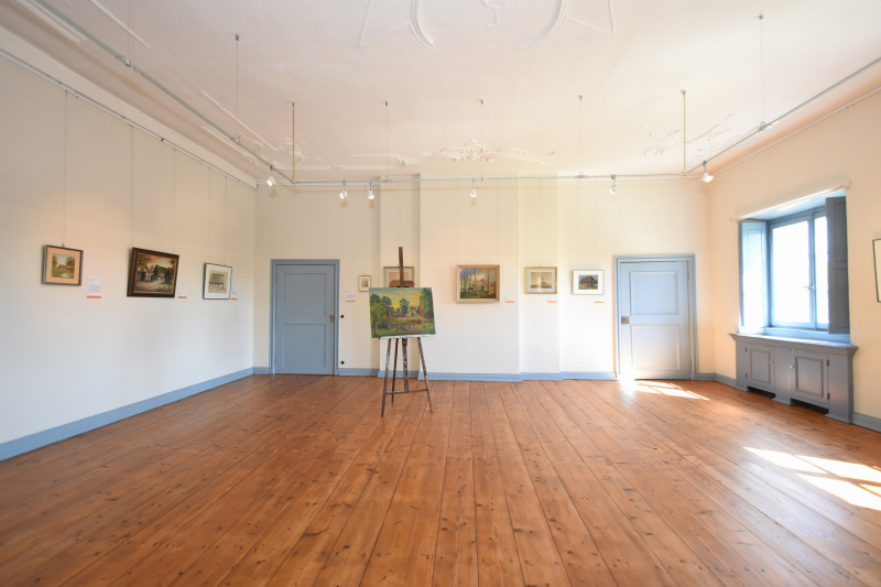 Einer der Ausstellungsräume mit hellem Holzboden, weißen Decken und blauen Fenstern und Türen