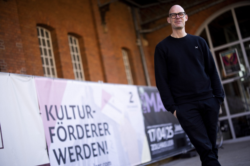 Mann mit Brille vor einem Banner mit dem Slogan "Kulturförderer werden!"