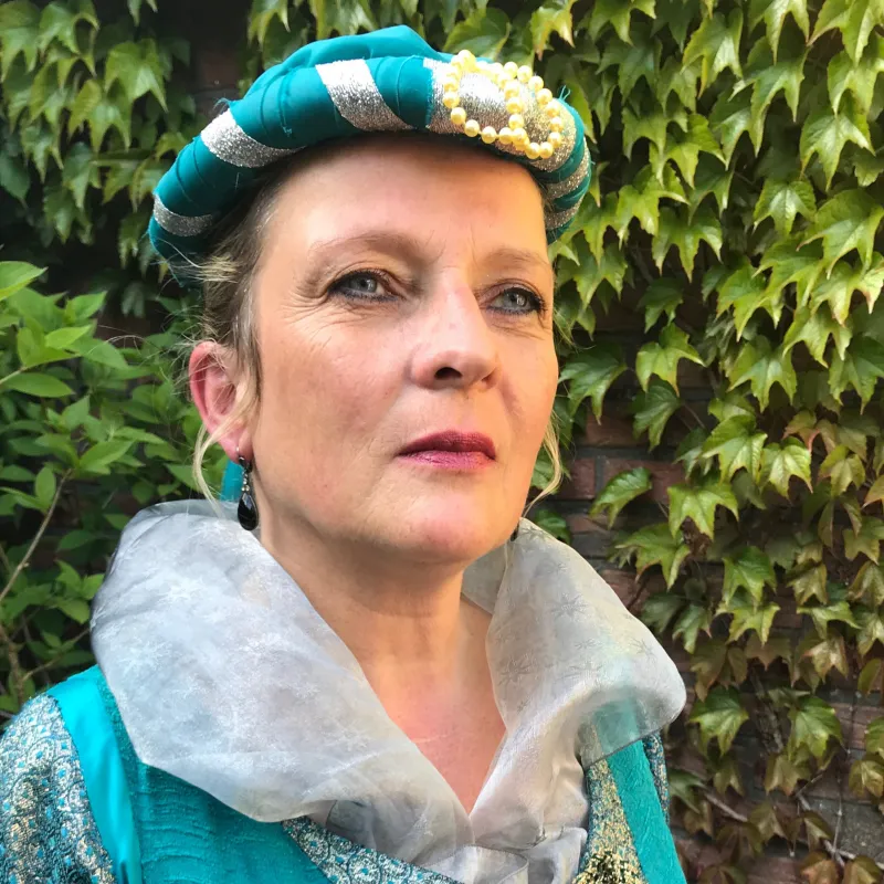Frau in historischem Kostüm mit türkis-silberner Haube und besticktem Kleid vor grüner Efeuwand.