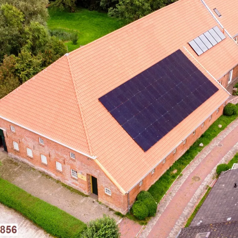 Backsteingebäude mit rotem Ziegeldach und Solaranlage, umgeben von Grünflächen und Pflasterweg.
