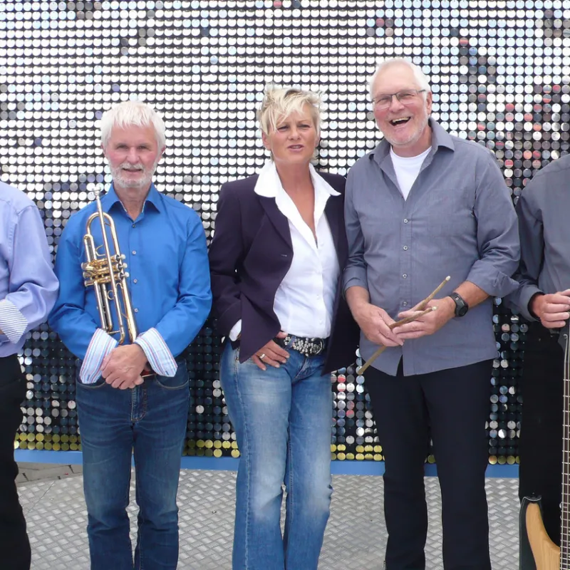 Fünf Personen stehen nebeneinander vor einer Wand mit reflektierenden runden Elementen, vier halten Musikinstrumente, eine Person trägt Jeans und ein weißes Hemd unter einer dunklen Jacke.