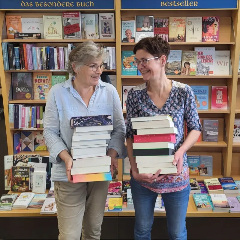 Zwei Frauen stehen vor Bücherregalen mit den Überschriften 'Das besondere Buch' und 'Bestseller' und halten jeweils einen großen Stapel Bücher.