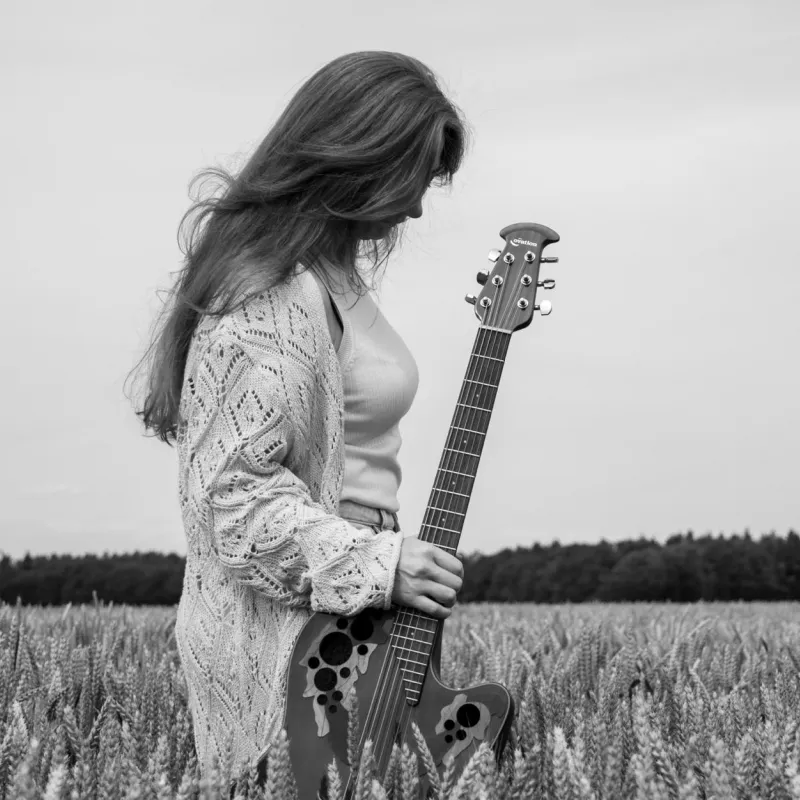 Frau mit langem Haar und Strickjacke steht in einem Getreidefeld und hält eine Akustikgitarre im Stehen