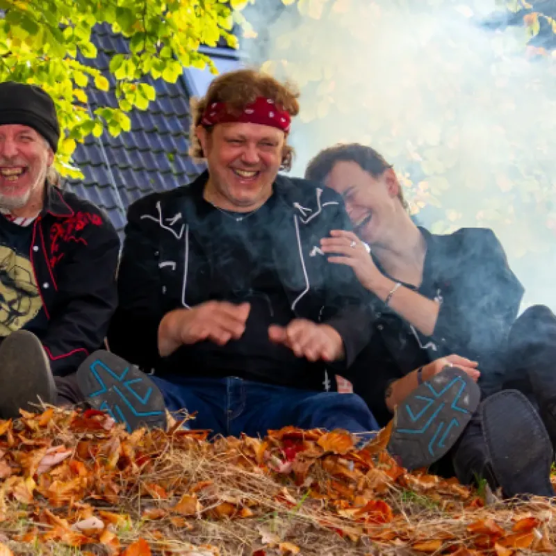 Drei Personen sitzen auf einem mit Herbstlaub bedeckten Boden vor einem Hausdach, umgeben von Rauch und Bäumen mit grünen und gelben Blättern