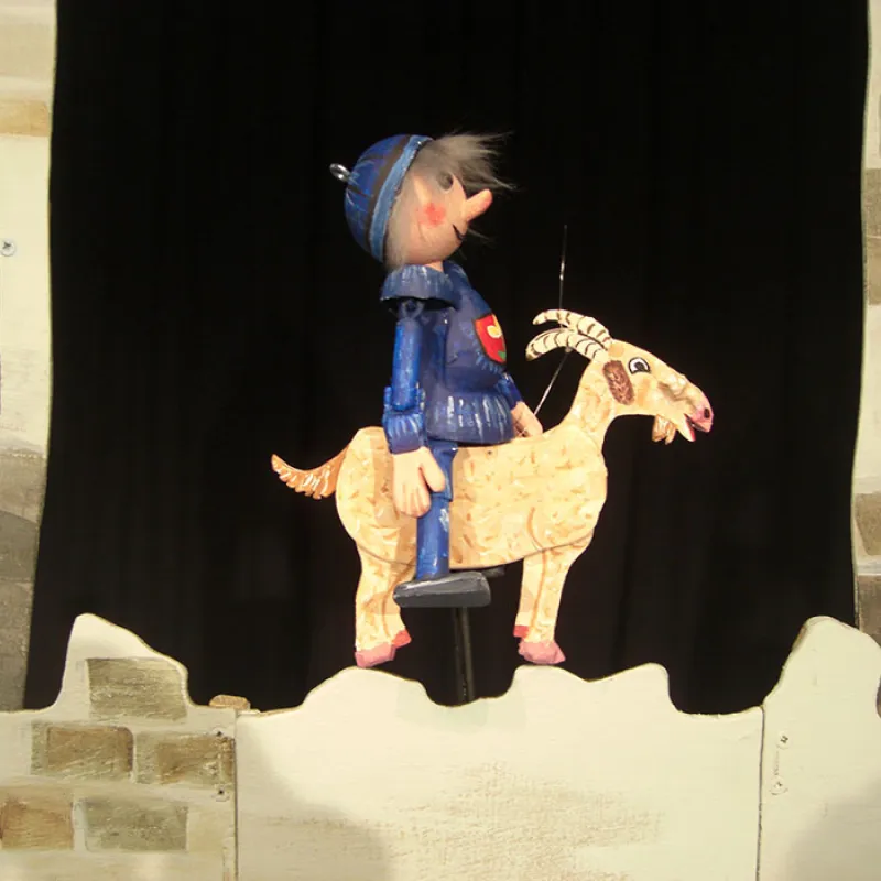 Puppentheater: Kleiner Ritter in blauer Rüstung reitet auf einer Ziege vor einer Burgmauer.