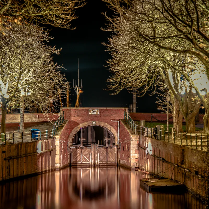 Beleuchtete Schleuse in Greetsiel, umgeben von kahlen Bäumen, spiegelt sich im ruhigen Wasser der Nacht.