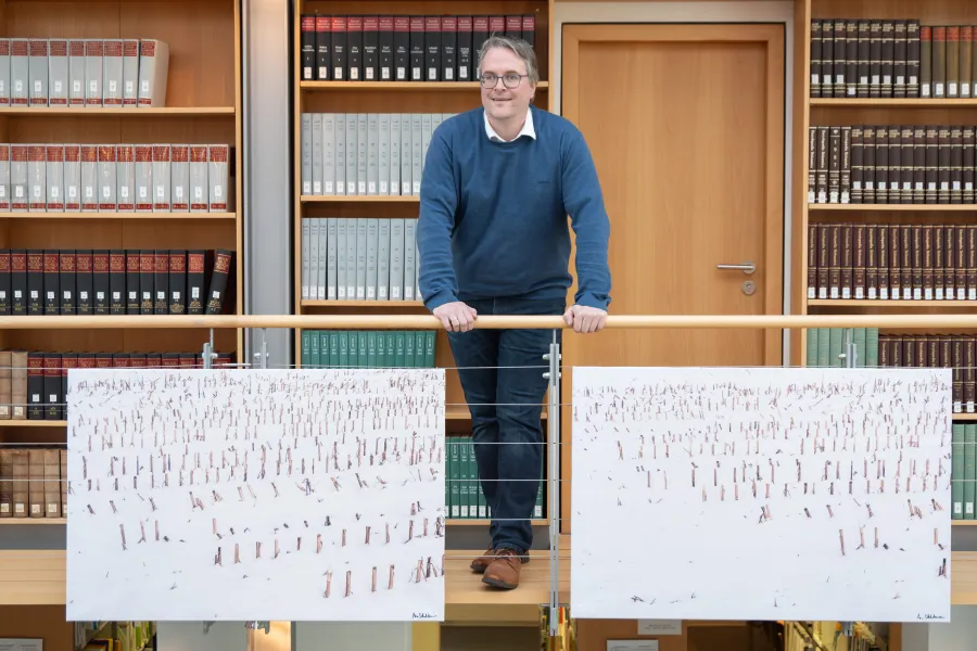 Dr. Heiko Suhr auf der Bibliotheksempore mit zwei Bildern, die senkrechte Strukturen zeigen, bei denen es sich um abgeernteten Mais handelt.