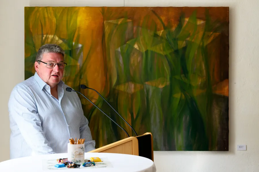 Gerald Hagen begrüßt Künstlerinnen und Gäste; im Hintergrund ein Gemälde von Gaby Mrongowius